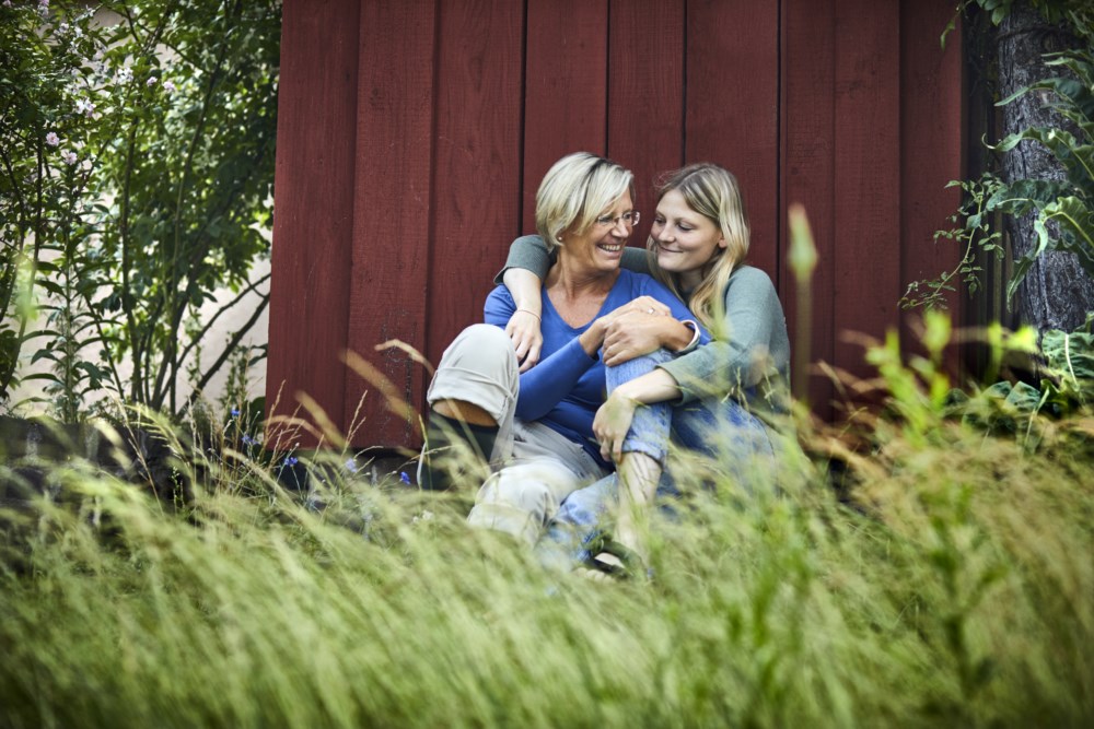Mor og datter som sitter i gresset utenfor rød hytte