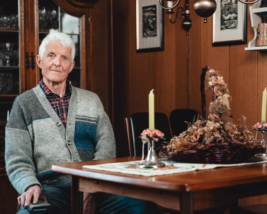 En eldre mann med hvitt hår og en grå-strikket jakke sitter ved et trebord i en tradisjonell norsk stue. Han smiler mildt mens han holder en liten bok i hånden. Bordet er pyntet med blomster og levende lys, og veggene i bakgrunnen er dekket av trepanel og dekorert med innrammede bilder. Atmosfæren er varm og hjemmekoselig.