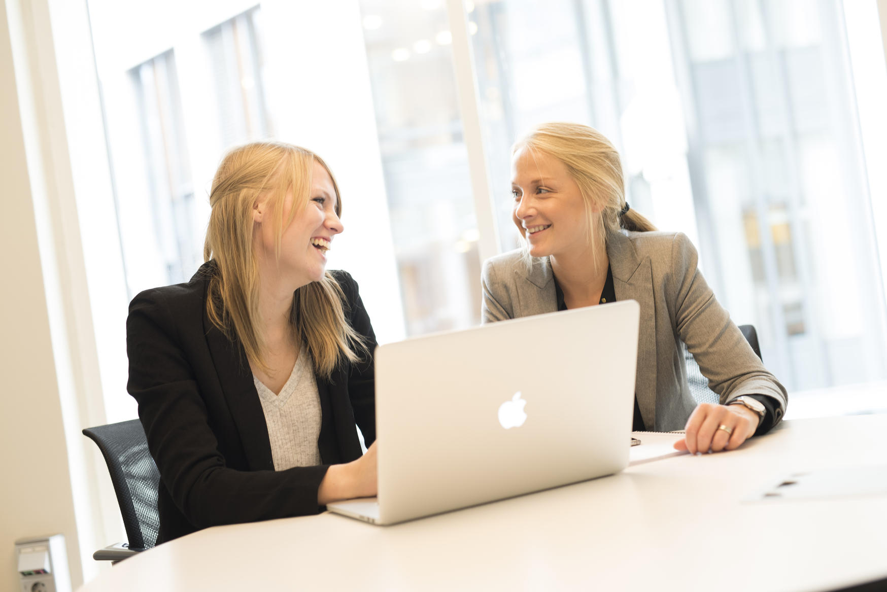 To damer med dressjakke, lyst hår og pc sitter ved kontorpult. foto